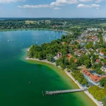 Luftbild von Herrsching am Ammersee: zeigt türkisgrünes Wasser, diverse Boote, einen langen Steg, eine bewaldete Landspitze und eine angrenzende Stadt mit dichter hellroter und weißer Bebauung. Rechts dahinter größere Gewässerflächen.