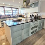 Das Bild zeigt eine hochwertige Einbauküche mit moderner Ausstattung. Die Küche hat weiße Schränke, eine zentrale Kochinsel mit eingebautem Herd und einer dunklen Arbeitsplatte. Links befindet sich ein großes Fenster mit Jalousien, rechts eine Sitzecke mit zwei grauen Sesseln und einem kleinen Tisch.