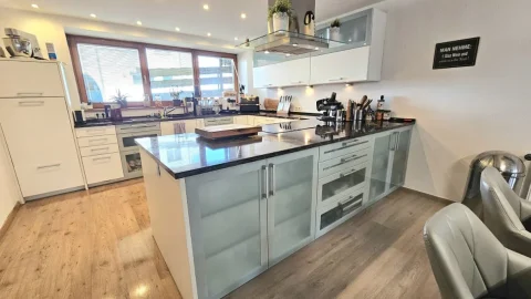 Das Bild zeigt eine hochwertige Einbauküche mit moderner Ausstattung. Die Küche hat weiße Schränke, eine zentrale Kochinsel mit eingebautem Herd und einer dunklen Arbeitsplatte. Links befindet sich ein großes Fenster mit Jalousien, rechts eine Sitzecke mit zwei grauen Sesseln und einem kleinen Tisch.