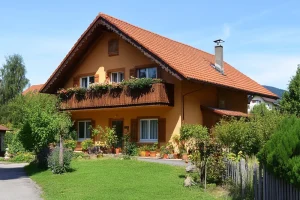 Ein großes, hochwertiges Einfamilienhaus in Wessling, Bayern, mit Satteldach und zwei Stockwerken. Die Fassade ist ockergelb mit weißen Fensterrahmen. Zwei Balkone mit Blumenkästen, umgeben von einem gepflegten Garten.