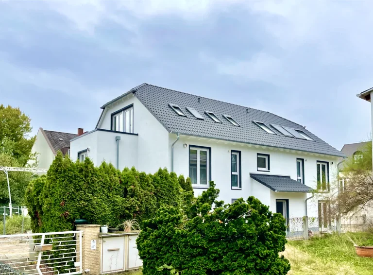 Ein weißes, modernes Reihenhaus unter einem grauen Dach. Am Dach befinden sich mehrere Dachfenster. Um das Haus sind Hecken und ein Metallzaun, dahinter ein blauer Himmel mit Wolken. Der String "IMG 0021 002" liefert keine zusätzlichen Informationen.