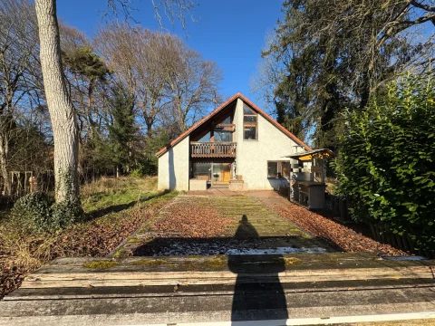 Auf dem Bild sieht man ein zweistöckiges Haus mit Satteldach und einem Balkon. Vor dem Haus befindet sich ein Garten mit Laub und Bäumen. Rechts steht eine dichte Hecke, links sieht man einen Baumstamm. Der Vordergrund zeigt einen alten Holzsteg.