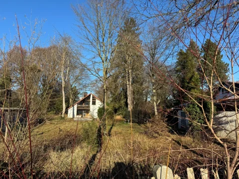 Das Bild zeigt ein ländliches Anwesen mit einem zweistöckigen Haus, das verwittert wirkt und von Bäumen sowie Büschen umgeben ist. Der Vordergrund ist mit wildem Gras und Gestrüpp bedeckt. Die natürliche Umgebung wirkt leicht verwildert und unbebaut.