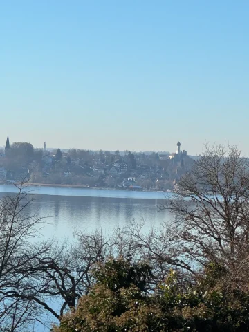 Das Bild zeigt eine Seenlandschaft mit einer Stadt am gegenüberliegenden Ufer. Im Vordergrund sind Bäume und Sträucher. Die Stadt umfasst Gebäude mit roten Dächern und hohen Strukturen wie Türmen. Gegenüber dem Wasser erscheint die Szene sonnig und ruhig.