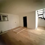 Ein leerer Raum mit Laminatboden und weiß gestrichenen Wänden. Links zwei kleine, rechteckige Fenster und eine schwarze Tür. Rechts eine Holztreppe, die nach oben führt. In der Mitte eine Metallschiene am Boden.