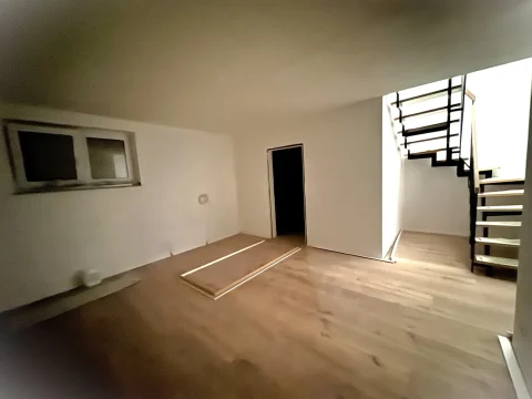 Ein leerer Raum mit Laminatboden und weiß gestrichenen Wänden. Links zwei kleine, rechteckige Fenster und eine schwarze Tür. Rechts eine Holztreppe, die nach oben führt. In der Mitte eine Metallschiene am Boden.