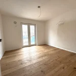 Das Bild zeigt ein leeres Zimmer mit hellen Wänden und einem dunkleren Holzboden. Es gibt eine Terrassentür, die nach draußen führt, und an der Decke sind offene Kabelenden für Leuchten zu sehen. Der Raum wirkt neu und unbewohnt.