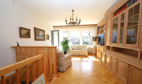 Wohnzimmer mit Parkettboden, beigen Sofas, einem Kronleuchter und einer Bücherwand. Drei Fenster bieten Aussicht auf Bäume. An den Wänden hängen Gemälde, und es gibt eine Holztreppe links.
