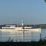 Das Bild zeigt ein großes, weißes Dampfschiff mit einem Schornstein und zwei Decks. Es fährt auf einem See, umgeben von einer hügeligen Landschaft. Menschen sind auf dem Schiff sichtbar, und es ist mit Girlanden geschmückt.