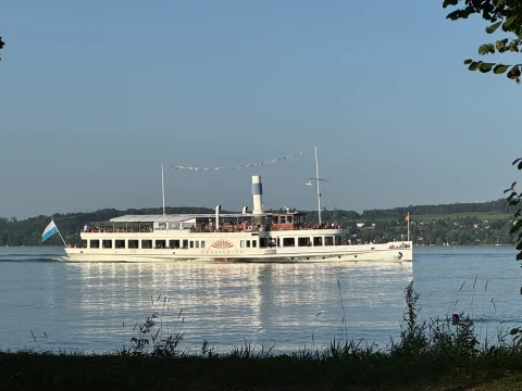 Der Dampfer Herrsching am Ammersee Das Bild zeigt ein großes, weißes Dampfschiff mit einem Schornstein und zwei Decks. Es fährt auf einem See, umgeben von einer hügeligen Landschaft. Menschen sind auf dem Schiff sichtbar, und es ist mit Girlanden geschmückt.
