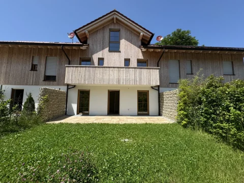 Das Bild zeigt ein modernes zweistöckiges Haus mit holzverkleideter Fassade. Das Erdgeschoss hat große Glasfenstertüren, und das Dach ist mit Satellitenschüsseln ausgestattet. Vor dem Haus ist eine Wiese und ein betonierter Patio.