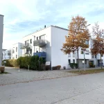 Auf dem Bild sind moderne, mehrstöckige Wohnhäuser in einer ruhigen Straße zu sehen. Die Gebäude sind weiß mit grauen Balkonen. Davor stehen zwei Bäume mit Herbstlaub. Der Himmel ist bewölkt. Es gibt Straßenpflaster und vereinzelt Fahrräder.