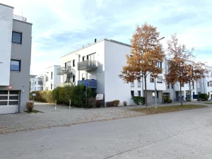 Auf dem Bild sind moderne, mehrstöckige Wohnhäuser in einer ruhigen Straße zu sehen. Die Gebäude sind weiß mit grauen Balkonen. Davor stehen zwei Bäume mit Herbstlaub. Der Himmel ist bewölkt. Es gibt Straßenpflaster und vereinzelt Fahrräder.