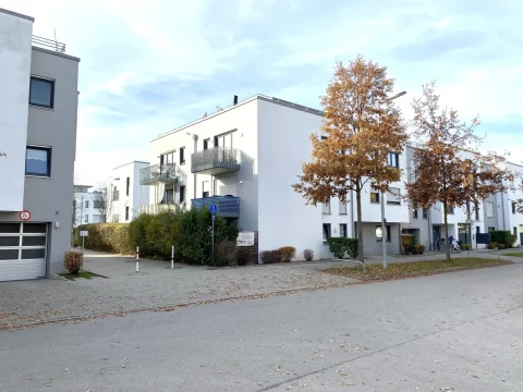 Wohnung Aubing zu verkaufen Auf dem Bild sind moderne, mehrstöckige Wohnhäuser in einer ruhigen Straße zu sehen. Die Gebäude sind weiß mit grauen Balkonen. Davor stehen zwei Bäume mit Herbstlaub. Der Himmel ist bewölkt. Es gibt Straßenpflaster und vereinzelt Fahrräder.