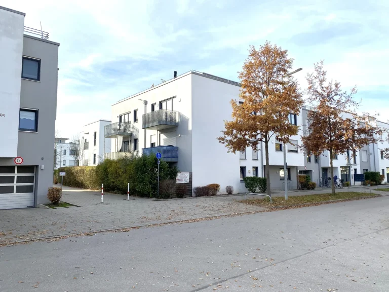 Auf dem Bild sind moderne, mehrstöckige Wohnhäuser in einer ruhigen Straße zu sehen. Die Gebäude sind weiß mit grauen Balkonen. Davor stehen zwei Bäume mit Herbstlaub. Der Himmel ist bewölkt. Es gibt Straßenpflaster und vereinzelt Fahrräder.