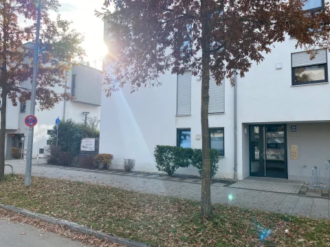 Eingangsbereich Das Bild zeigt ein modernes, weißes Mehrfamilienhaus mit großen Fenstern und zwei Eingängen. Vor dem Haus stehen Herbstbäume mit braunen Blättern und ein Verkehrsschild, das das Halten verbietet. Die Sonne scheint zwischen den Ästen durch.