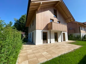 Auf dem Foto ist ein modernes zweistöckiges Haus mit hölzerner Fassade und weißen Wänden zu sehen. Es hat eine große Terrasse mit Steinfliesen und einen gepflegten Rasen. Ideal für Immobilienmakler am Ammersee Süd.