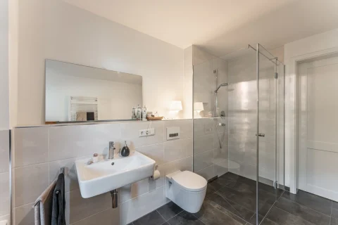 Helles, modernes Badezimmer mit großem Spiegel, Waschbecken, Toilette und gläserner Duschkabine. Graue Fliesen vervollständigen den eleganten Look, ideal für Immobilienangebote in Schliersee.