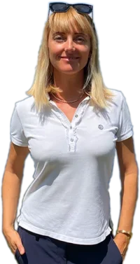 Auf dem Bild Myriam Goldhofer (Immobilienmakler am Ammersee) in einem weißen Poloshirt und dunkler Hose zu sehen. Sie trägt eine Sonnenbrille auf dem Kopf. Ihr Lächeln und ihre entspannte Haltung lassen sie freundlich und professionell wirken. Ihr Outfit ist leger und sommerlich.