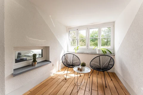 Helle Innenloggia mit modernem Design und Grünblick Das Bild zeigt eine Innenloggia mit schrägen Wänden und Fenstern mit Blick auf grüne Bäume. Ein Holzboden und zwei moderne schwarze Drahtstühle um einen weißen Rundtisch sind zentral platziert. An der Wand ist ein Einbau-Kamin.