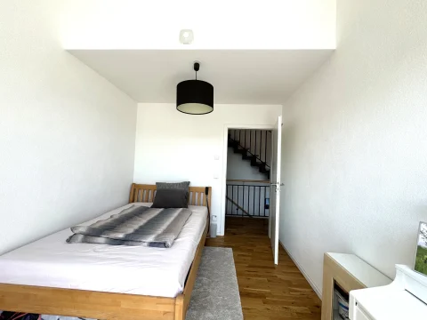 Das Bild zeigt ein minimalistisch eingerichtetes Zimmer mit einem Einzelbett, einer schwarzen Pendelleuchte und einem offenen Türrahmen, der zu einer Treppe führt. Ein kleiner, weißer Tisch steht rechts im Bild.