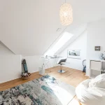 Das Bild zeigt ein helles Dachgeschoss, das als multifunktionales Kinderzimmer und Büro genutzt wird. Es enthält eine Ecke mit einem Schreibtisch und Stuhl, eine Skaterboard, eine runde Sitzgelegenheit sowie eine Spielfläche mit Teppich.