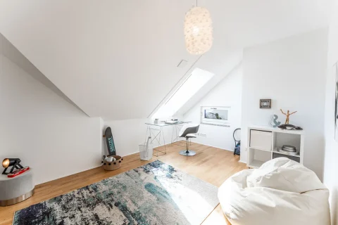 Modernes Kinderzimmer und Büro unter einem Dach Das Bild zeigt ein helles Dachgeschoss, das als multifunktionales Kinderzimmer und Büro genutzt wird. Es enthält eine Ecke mit einem Schreibtisch und Stuhl, eine Skaterboard, eine runde Sitzgelegenheit sowie eine Spielfläche mit Teppich.