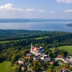 Dieses Luftbild zeigt das Kloster Andechs in ländlicher Umgebung oberhalb des Ammersees. Das barocke Ensemble wird umgeben von Wäldern und landwirtschaftlichen Flächen, während im Hintergrund der weite, blau glitzernde Ammersee und umliegende Ortschaften zu sehen sind.