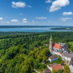 Luftaufnahme des Klosters Andechs in Bayern, umgeben von üppigen Wäldern und Wiesen, mit Blick auf den Ammersee. Die Architektur umfasst eine markante rote Dachkirche. Ein idealer Ort für Immobilieninteressierte.