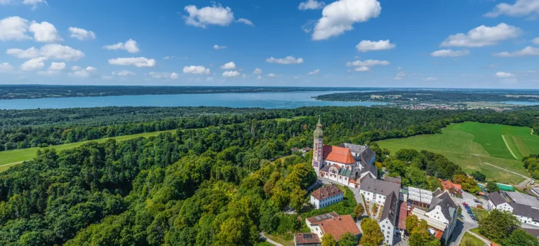 Luftaufnahme des Klosters Andechs in Bayern, umgeben von üppigen Wäldern und Wiesen, mit Blick auf den Ammersee. Die Architektur umfasst eine markante rote Dachkirche. Ein idealer Ort für Immobilieninteressierte.