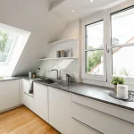 Ein helles Dachgeschoss-Kücheninterieur mit modernen, weißen Schränken und grauer Arbeitsplatte. Zwei Fenster sorgen für Tageslicht und Ausblick auf Bäume. Zubehör fürs Kochen ist vorhanden, inklusive Spüle und Armaturen. Dekorative Elemente wie Pflanzen und ein Bild zieren den Raum.