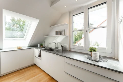 Helle Küche unter Dachfenstern – Perfekt zum Kochen Ein helles Dachgeschoss-Kücheninterieur mit modernen, weißen Schränken und grauer Arbeitsplatte. Zwei Fenster sorgen für Tageslicht und Ausblick auf Bäume. Zubehör fürs Kochen ist vorhanden, inklusive Spüle und Armaturen. Dekorative Elemente wie Pflanzen und ein Bild zieren den Raum.