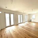 Ein leerer Wohnraum mit Holzfußboden, mehreren Fenstertüren zur Terrasse und einer Fensterseite. Unmöbliert, ideal für individuelle Gestaltung mit Koch-, Ess- und Wohnbereich.