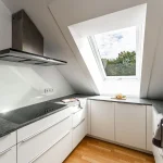 Eine elegante, moderne Küche unter einer Dachschräge mit einem großen Fenster, das den Raum erhellt. Die Küche ist mit weißen Schränken und einer dunklen Marmorarbeitsplatte ausgestattet. Eine innovative Dunstabzugshaube und eine integrierte Kochplatte vervollständigen das Bild des Kochens mit Spaß.