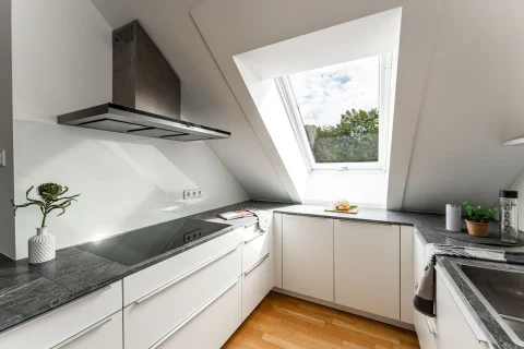 Moderne Küche unter Dachschräge – Kochen mit Spaß Eine elegante, moderne Küche unter einer Dachschräge mit einem großen Fenster, das den Raum erhellt. Die Küche ist mit weißen Schränken und einer dunklen Marmorarbeitsplatte ausgestattet. Eine innovative Dunstabzugshaube und eine integrierte Kochplatte vervollständigen das Bild des Kochens mit Spaß.