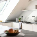 Moderne Dachgeschossküche mit hellen Schränken, grauer Arbeitsplatte, Einbauspüle und stilvollen Deko-Elementen. Ein Dachfenster sorgt für natürliches Licht.
