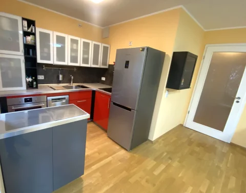 Geräumige Küche mit moderner Ausstattung und Flurblick Die Küche zeigt moderne Einrichtung mit einer Kombination aus weißen Oberschränken und roten Unterschränken. In der Mitte steht eine graue Kochinsel. Rechts im Bild ist eine Tür, die vermutlich zum Flur führt. Ein großer Kühlschrank steht neben einem schwarzen Wandregal.