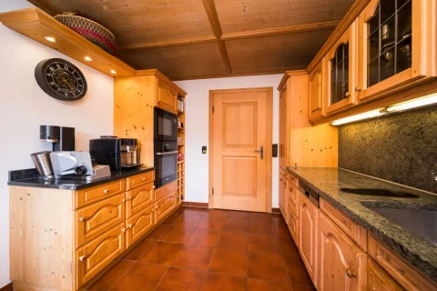 Einbauküche im Tegernseer Landhausstil Rustikale Küche in einem Haus am Tegernsee mit holzvertäfelten Wänden und Decke, Holzschränken, dunklem Granitarbeitsplatten, modernen Küchengeräten und einer runden Wanduhr.