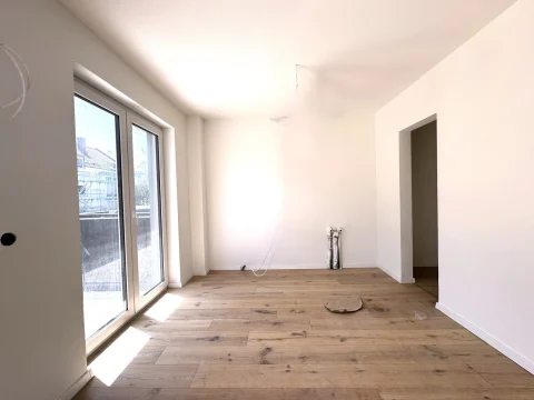 Hier ist ein Raum mit großen Fenstern und Holzboden sichtbar. Rechts hinten ist eine Türöffnung. Vorne stehen Wasseranschlüsse, möglicherweise für einen Küchenbereich bestimmt. Der Raum scheint renoviert oder neu gebaut zu sein.