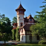 Das Bild zeigt das Kurparkschlösschen in Herrsching am Ammersee, ein malerisches Bauwerk im Fachwerkstil mit einem markanten Turm, umgeben von üppigem Grün und einem gepflegten Garten.