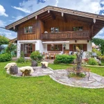 Das Foto zeigt ein charmantes Landhaus am Tegernsee mit einem gepflegten Garten. Der zweistöckige Holzbau hat eine große Terrasse und Balkone. Im Vorgarten sieht man eine Wiese, Blumenbeete, Hochbeete und eine Steinskulptur.