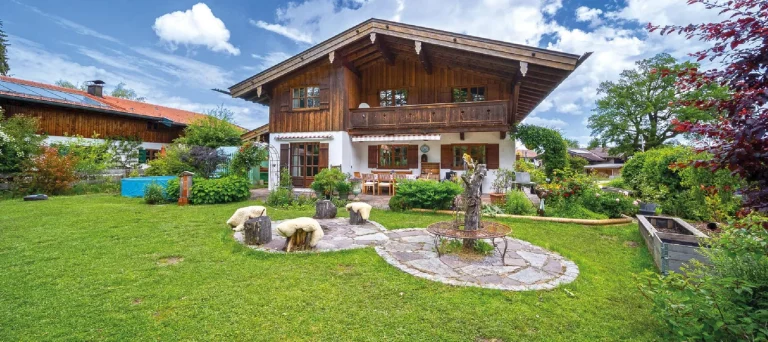Das Foto zeigt ein charmantes Landhaus am Tegernsee mit einem gepflegten Garten. Der zweistöckige Holzbau hat eine große Terrasse und Balkone. Im Vorgarten sieht man eine Wiese, Blumenbeete, Hochbeete und eine Steinskulptur.