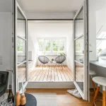 Das Bild zeigt eine offene Loggia mit Holzboden, zugänglich durch eine Schiebetür. Zwei moderne schwarzgestellte Stühle stehen um einen Beistelltisch, hinter dem ein großes Fenster ist. Rechts befindet sich eine Küchenzeile.