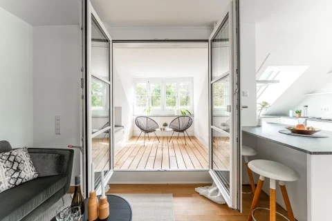 Moderne Loggia: Ein heller, friedvoller Rückzugsort Das Bild zeigt eine offene Loggia mit Holzboden, zugänglich durch eine Schiebetür. Zwei moderne schwarzgestellte Stühle stehen um einen Beistelltisch, hinter dem ein großes Fenster ist. Rechts befindet sich eine Küchenzeile.