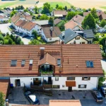 Luftaufnahme einer ländlichen Siedlung. Im Vordergrund ein großes Haus mit Ziegeldach und Dachfenstern. Umgeben von weiteren Wohnhäusern mit ähnlicher Architektur, Autos und sattgrüner Vegetation. Im Hintergrund Felder.