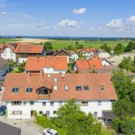 Luftaufnahme einer ländlichen Siedlung zeigt Einfamilienhäuser mit Ziegeldächern, einige mit Solaranlagen. Im Hintergrund Felder und eine Kirche. Helle Fassaden, grüne Gärten und Fahrzeuge sind sichtbar.