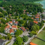 Luftbild von Schondorf am Ammersee zeigt eine idyllische Ortschaft mit dichter Bebauung, umgeben von Wald und direkt am Ufer des Ammersees. Man sieht Wohnhäuser, grüne Flächen und einen kleinen Bootshafen.