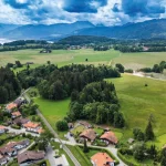 Das Luftbild zeigt eine ländliche Region am Tegernsee mit einer Siedlung von Häusern, umgeben von Wäldern und Feldern. Im Hintergrund sind Berge und der teilweise sichtbare Tegernsee zu erkennen.
