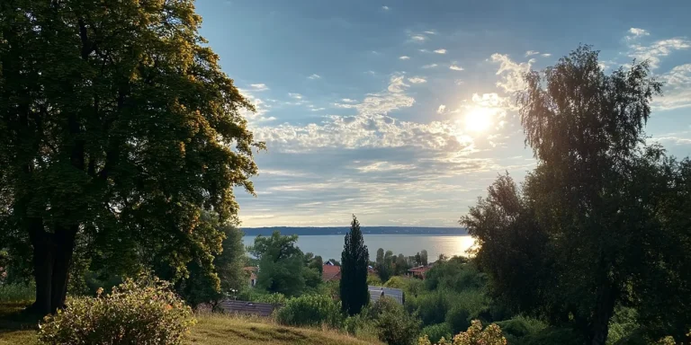 Das Bild zeigt eine malerische Aussicht mit einem Blick auf einen See, umrahmt von üppigen Bäumen unter einem dramatischen Himmel mit abendlichem Sonnenlicht. Im Vordergrund sind grüne Wiesen und einige Häuser erkennbar.