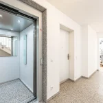 Ein moderner Aufzug mit Glasfront und Edelstahlrahmen, geöffnet im Erdgeschoss eines Gebäudes, sichtbar auf Granitboden. Nebenan eine geschlossene weiße Tür. Links führt ein Durchgang zu einem hellerleuchteten Raum mit Treppe.